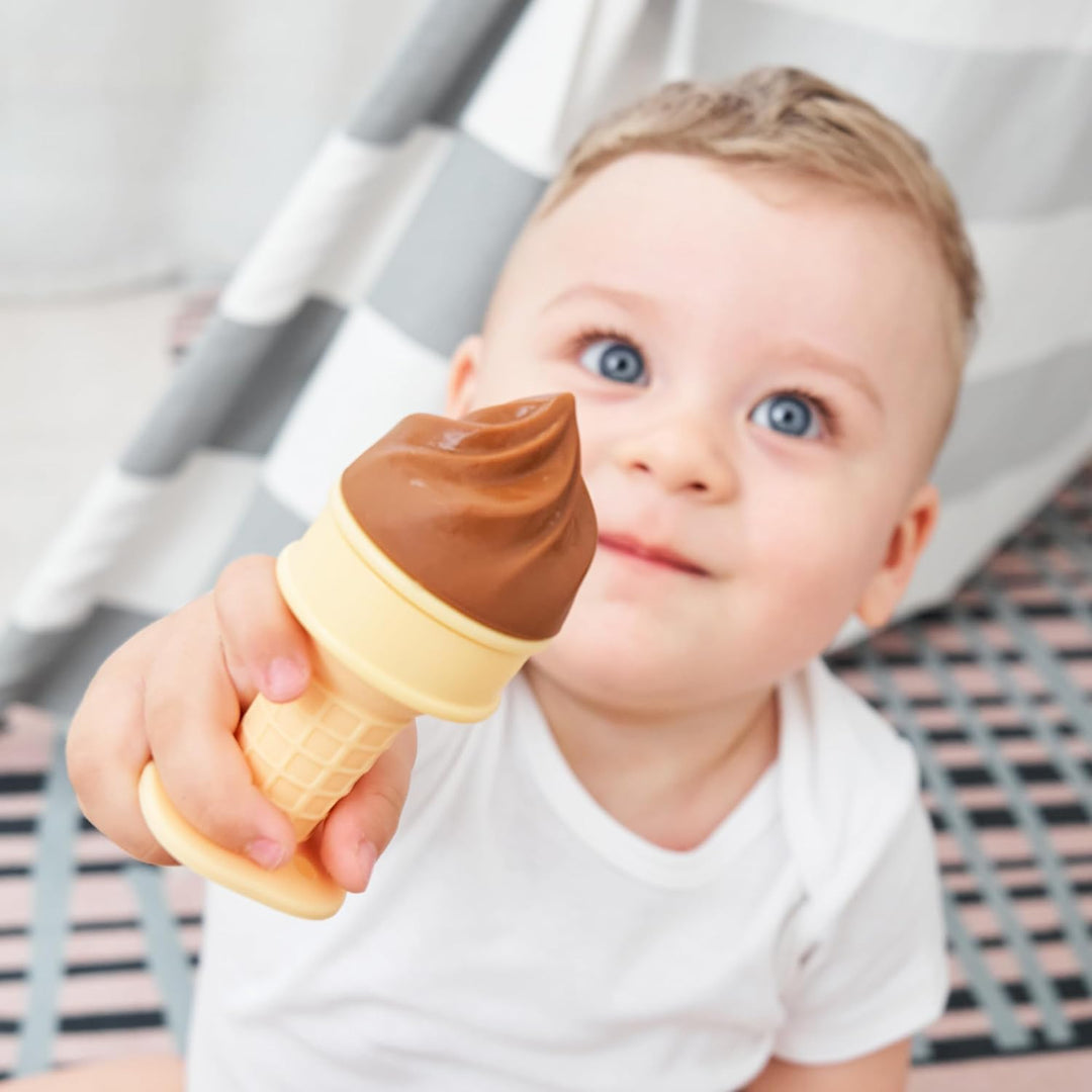 Happy blue-eyed baby holding up Smily Mia chocolate brown ice cream freezable teether toward camera showing easy palm grip and delighted expression