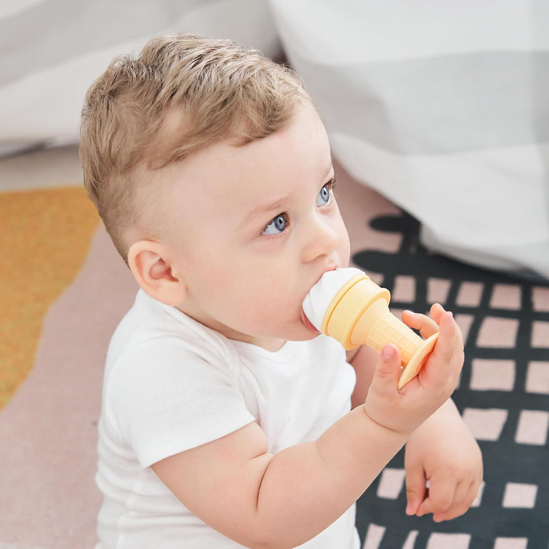 Blue-eyed baby sitting on floor confidently holding complete Smily Mia white ice cream freezable teether upright demonstrating easy grip cone handle for babies 4 months plus