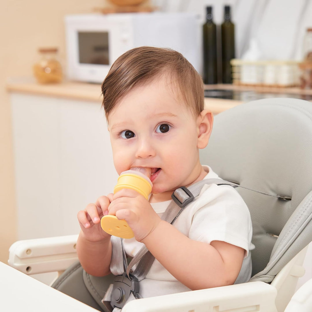 Happy baby sitting in high chair independently holding and chewing Smily Mia yellow ice cream cone teether for self-feeding and teething relief