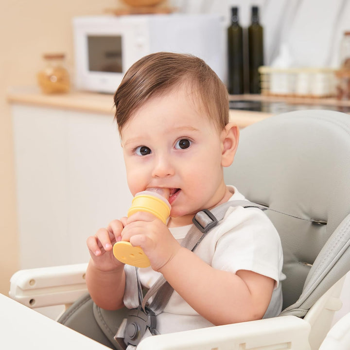 Happy baby sitting in high chair independently holding and chewing Smily Mia yellow ice cream cone teether for self-feeding and teething relief