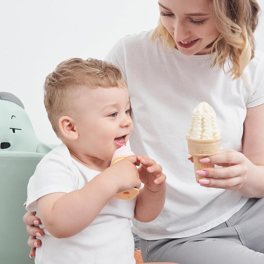 Toddler boy laughing while biting Smily Mia pink ice cream freezable teether as mother holds a matching real soft-serve cone beside him